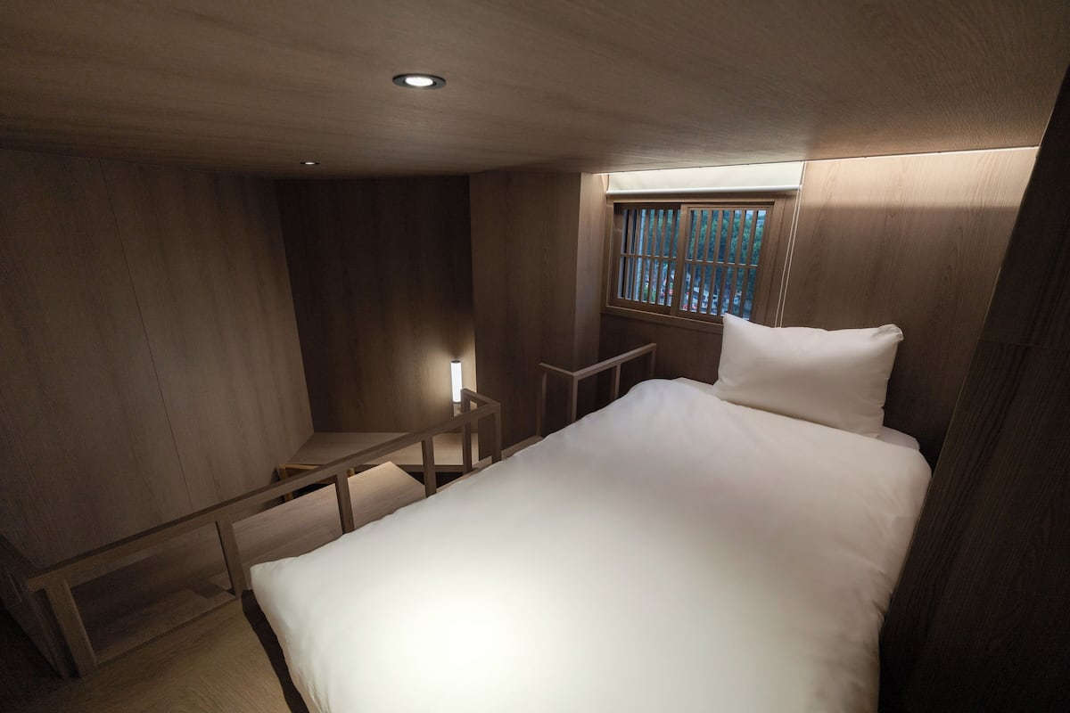 A bedroom with a bed, a nightstand, a window with bars, and a wooden ceiling.