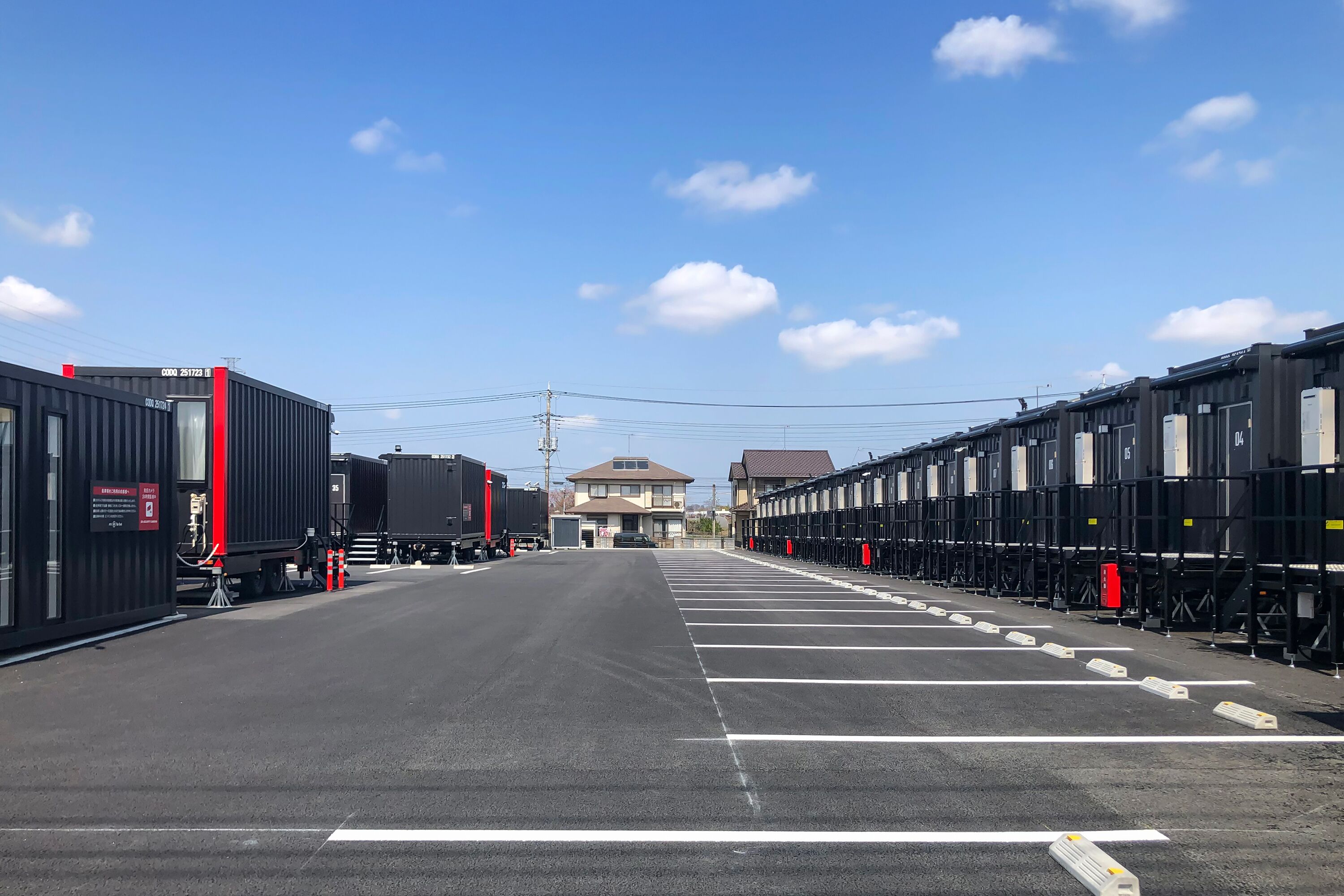 A parking area with rows of shipping containers, a building in the background, and a clear sky.