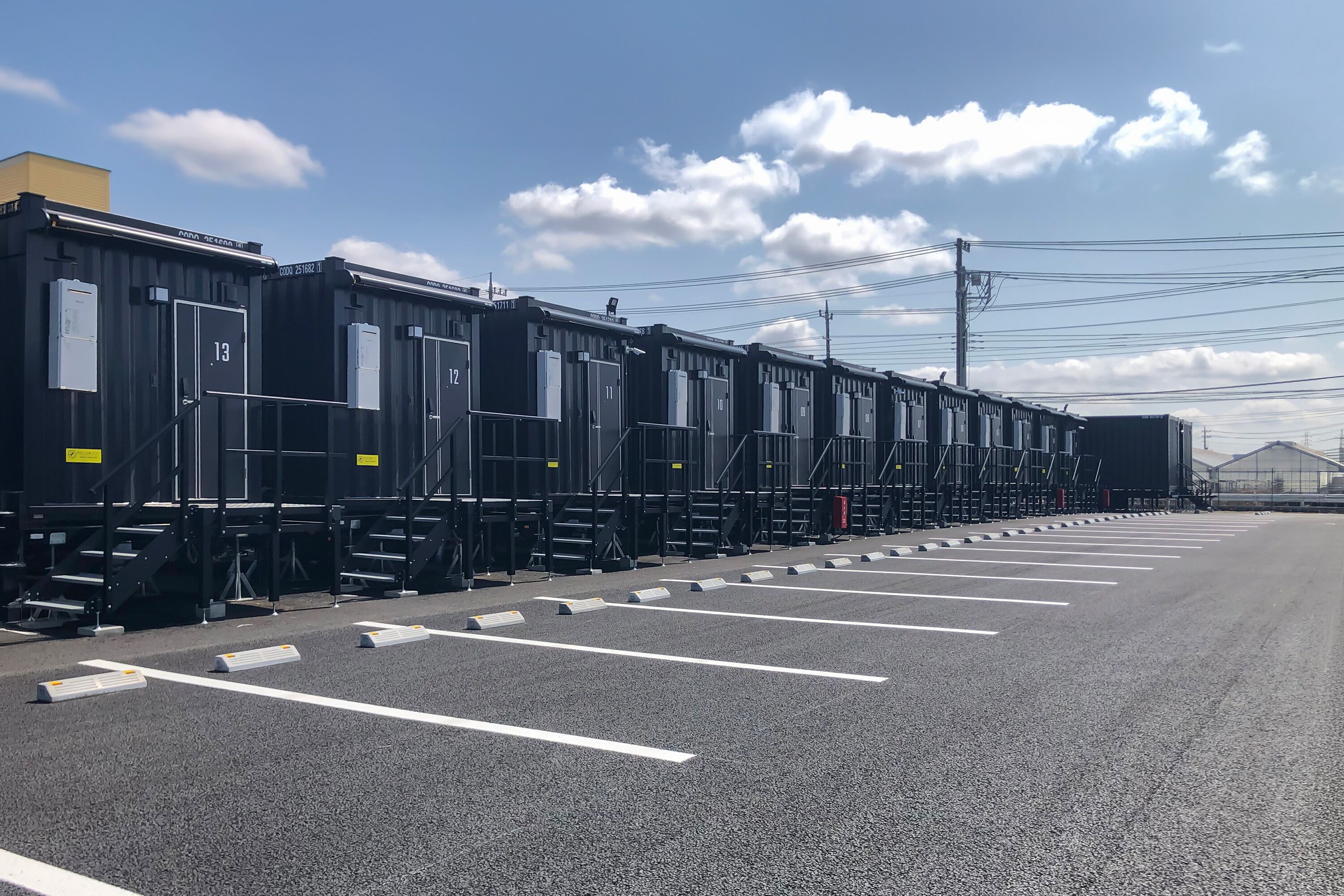 Fila de habitaciones de hotel modulares negras con puertas numeradas, un estacionamiento con espacios delimitados y un cielo despejado.