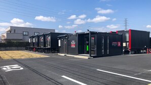 A parking lot with a row of black shipping containers, a building with a sign, and a clear blue sky. - HOTEL R9 The Yard Hanazonointer (Fukaya)