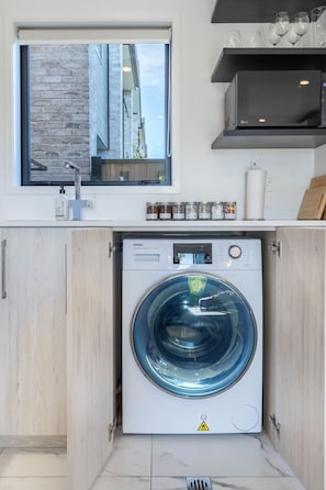 A compact laundry room with a front-loading washer and dryer, a window above the sink, and a small shelf with glassware. - Central Townhouse Near One NZ Stadium & Te Pae (Christchurch)