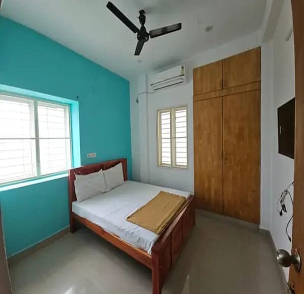 A bedroom with a bed, a ceiling fan, two windows, an air conditioning unit, and a wardrobe.