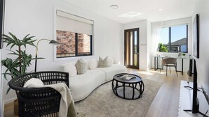 Un salon moderne avec un canapé blanc, un fauteuil en osier noir, une table basse ronde et un mur décoré d’un motif en briques.