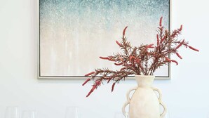 Une table à manger dressée avec de l’assiette, des bols et des verres blancs, un vase blanc contenant des feuilles rouges, et un tableau abstrait encadré accroché au mur.