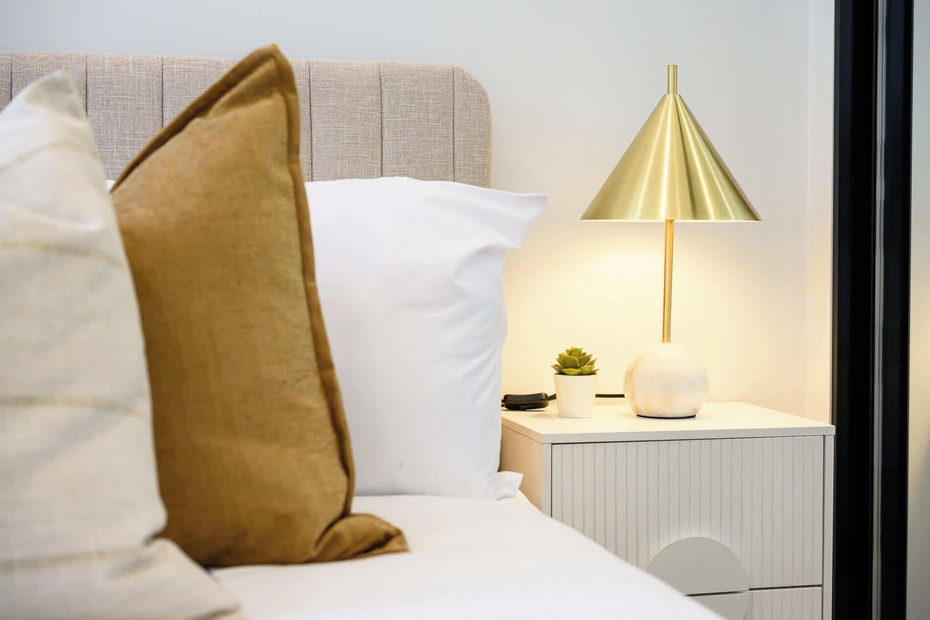 A neatly made bed with white and brown pillows, a bedside lamp, a small potted plant, and a white nightstand with a drawer.