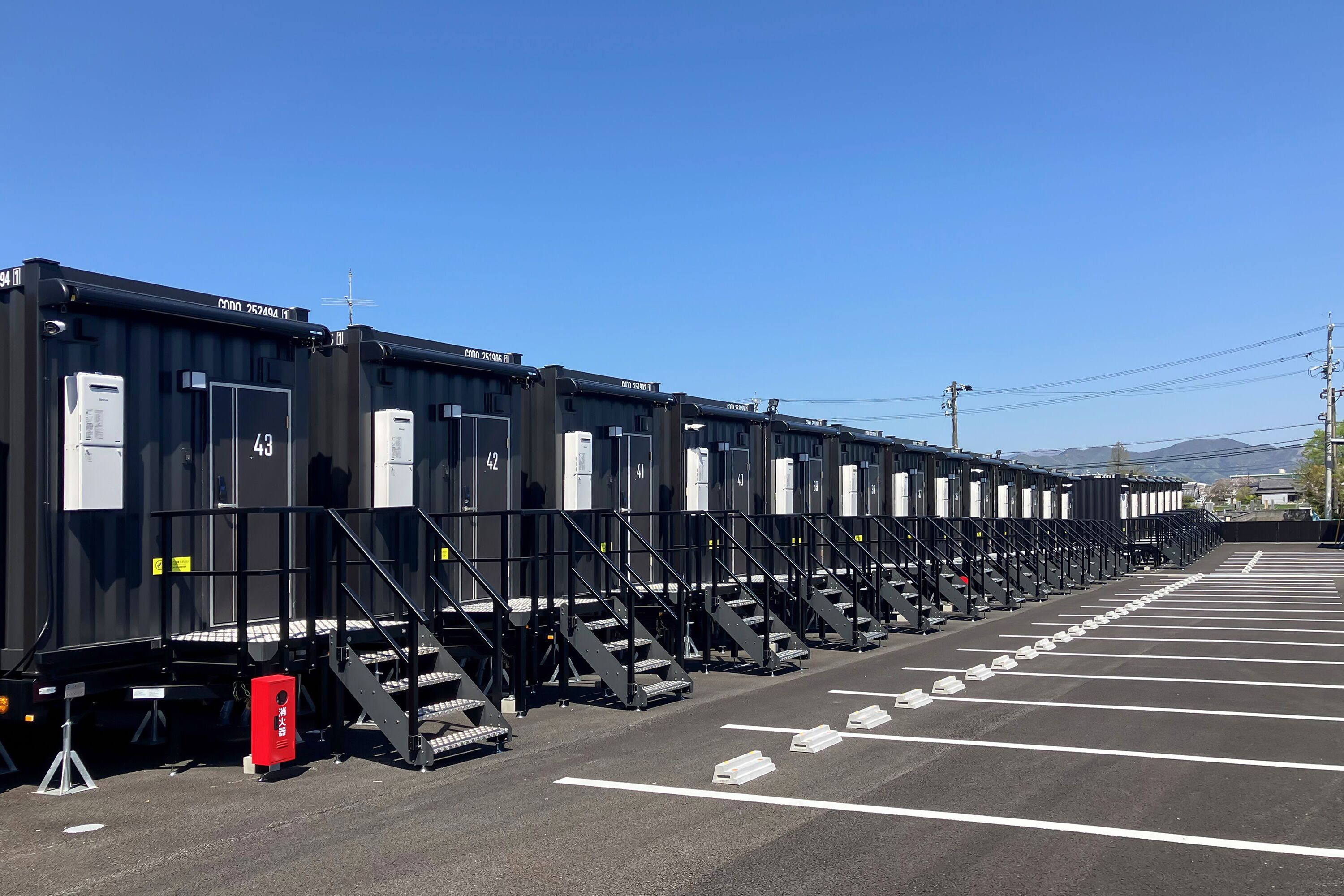 A row of black shipping containers with white text and numbers, arranged in a parking lot with white lines on the ground.