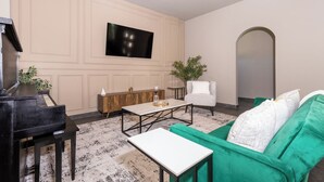 Een woonkamer met een piano, een aan de muur bevestigde televisie, een plafondventilator, een groene bank en een kleine tafel.