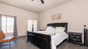 Een slaapkamer met een bed, een stoel, een nachtkastje, een dressoir, een plafondventilator en een raam met gordijnen.