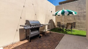 Een terras buiten met een barbecue, een tafel en stoelen, en een groen-witte parasol.