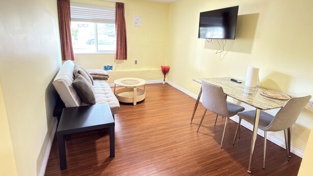Een woonkamer met een bank, een eettafel met stoelen, een flatscreen televisie aan de wand en een raam met gordijnen.