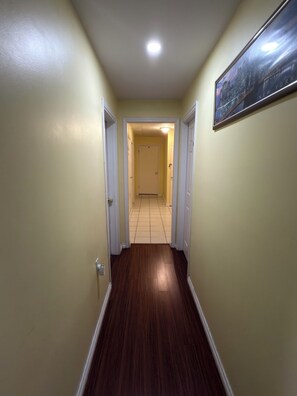 Un pasillo con piso de madera, paredes color crema y una puerta al final que da a una habitación con piso de baldosas.