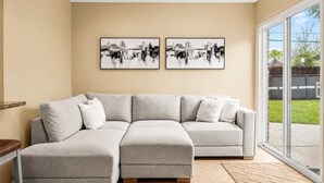 A living room with a sectional sofa, two framed abstract paintings, and a large sliding glass door leading to a backyard. - Private Pool Oasis in Quiet Energy Corridor Villa (Houston)