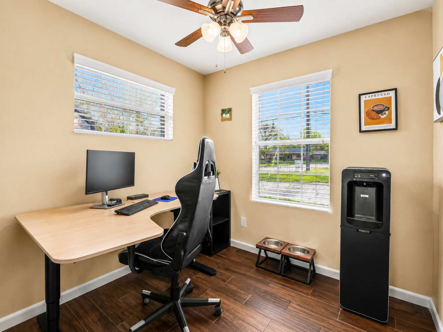 A home office with a desk, computer, chair, two windows, a ceiling fan, and a water dispenser.