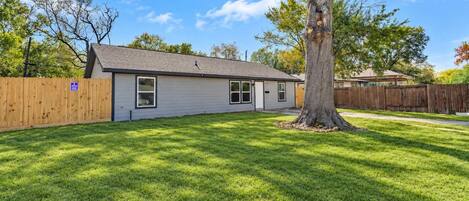 A single-story house with a fenced yard, a large tree, and a clear sky.