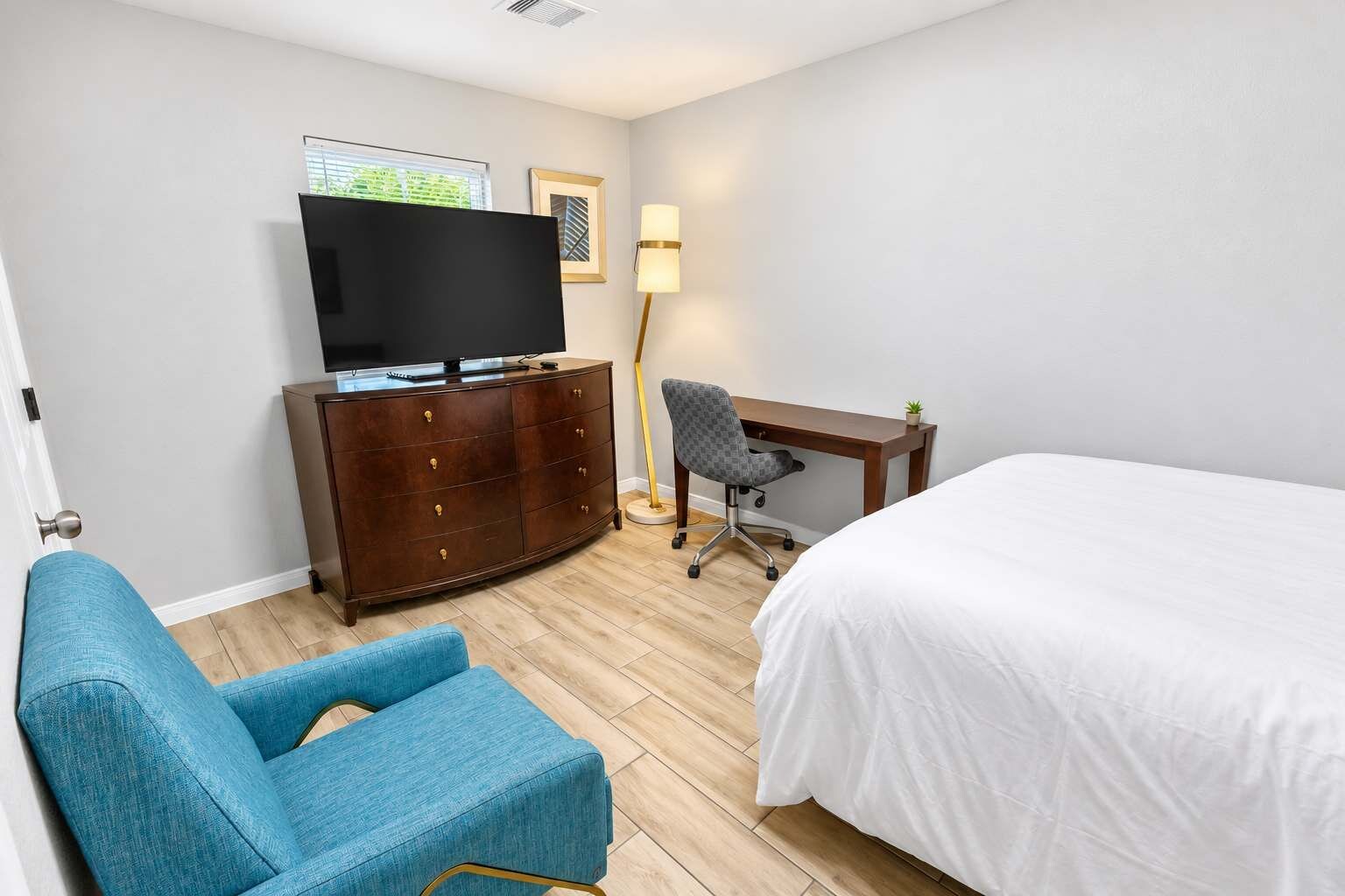 Una habitación de hotel con una cama, un escritorio, una silla, un televisor sobre un aparador y una lámpara.