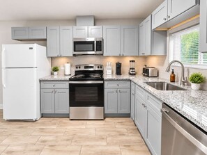 A modern kitchen with grey cabinets, stainless steel appliances, a granite countertop, and a window with blinds. - 4BR Crew Home | 2.5 Baths, 4 Desks, Sleeps 8 (Houston)