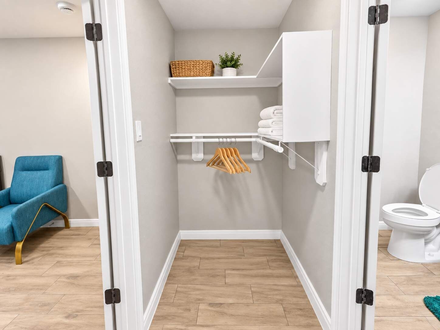 A bathroom with a white toilet, a shelf with folded towels, a hanging rack with towels, and a blue chair in the adjacent room.
