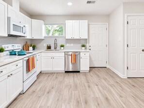 A modern kitchen with white cabinetry, a stainless steel dishwasher, and a window with blinds. - 4BR Crew Home | 4 Desks, Driveway, Near Hobby (Houston)