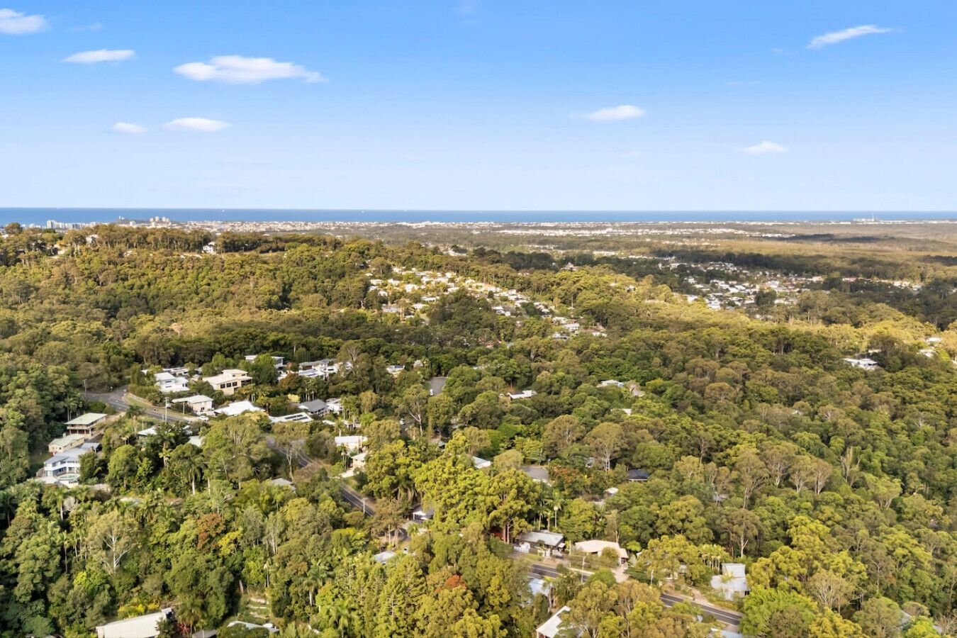 Une vue aérienne d’un quartier résidentiel, avec des maisons dispersées au milieu d’une végétation luxuriante, un ciel dégagé et une côte lointaine.