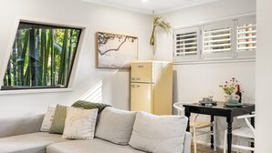 A modern living room with a sofa, a vintage refrigerator, a dining table with chairs, and a window with a view of bamboo.