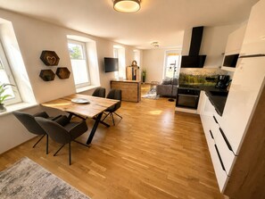 Eine moderne Küche mit Backofen, ein Essbereich mit Holztisch und -stühlen sowie ein Wohnbereich mit Sofa und Fernseher.