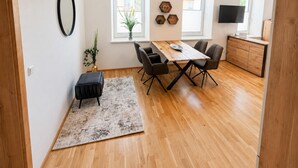 Une cuisine moderne avec des meubles en bois, une table à manger avec des chaises, un téléviseur noir fixé au mur et une fenêtre agrémentée d’une plante.