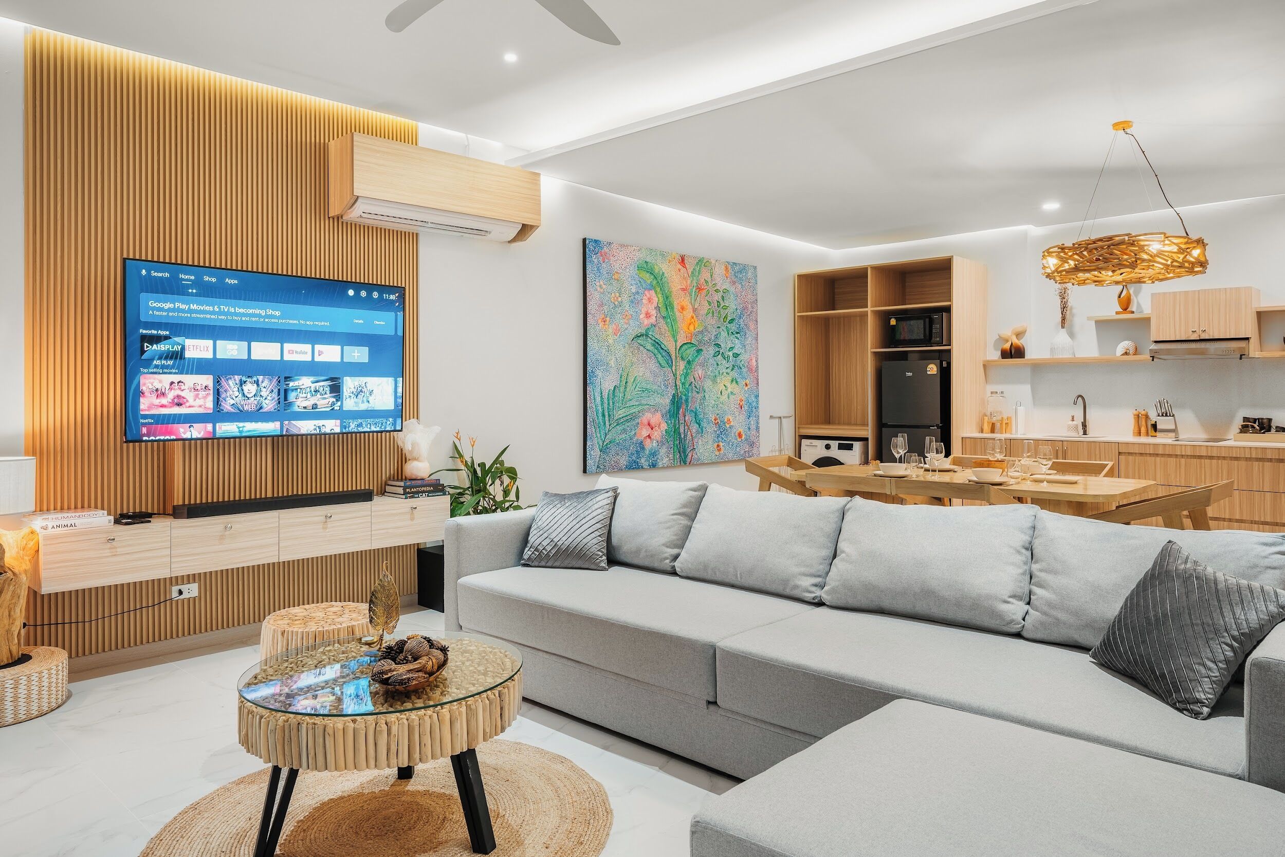 A modern living room with a flat-screen TV, a sofa, a dining table, and a kitchen area.