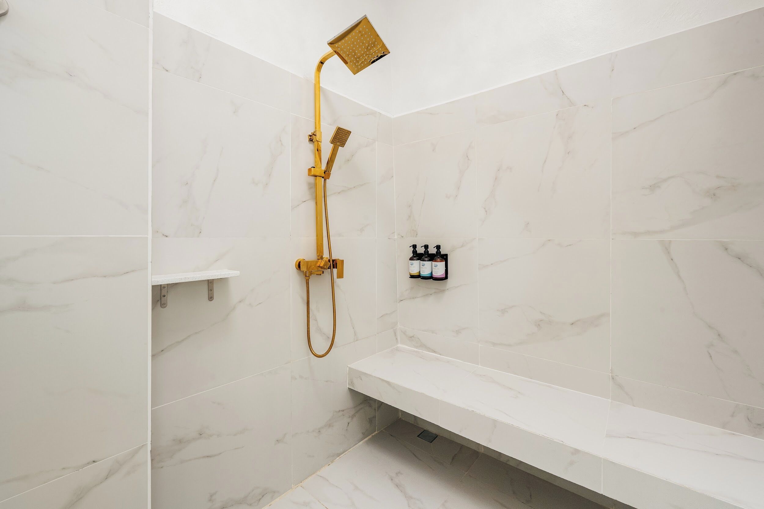 A bathroom with white marble walls, a gold showerhead, and a white bench.