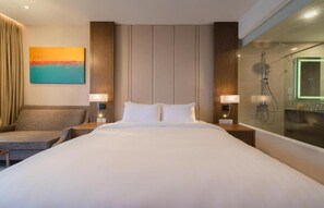 Ein Hotelzimmer mit einem großen Bett, zwei Nachttischen inklusive Lampen, einem Sofa, einem Bild an der Wand und einer Duschkabine aus Glas.