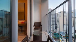 A balcony with a wicker chair and table, a view of tall buildings, and a door leading to a room with a desk and chair. - Panorama Luxury Hotel - Full Services (Nha Trang)