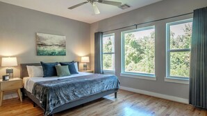 Una camera da letto con un letto, due comodini, una ventola a soffitto, ampie finestre e vista sugli alberi.