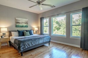 A bedroom with a bed, two bedside tables, a ceiling fan, large windows, and a view of trees. - Your Comfy Bungalows! Sleeps 30/12BR/8BA/16PA (Houston)