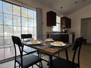 Uma área de refeições com uma mesa posta para quatro pessoas, uma cozinha ao fundo e vista através de amplas janelas.