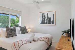 Een slaapkamer met een bed, kussens, een nachtkastje, een televisie, een plafondventilator en een ingelijste foto aan de muur.
