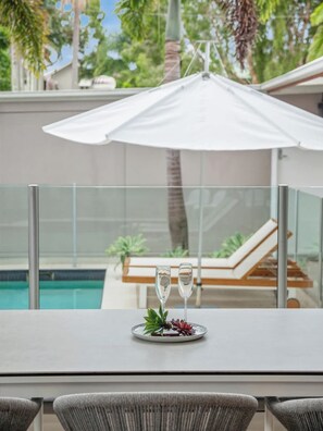 Een tafel met twee glazen en een bord eten, een parasol en een zwembad op de achtergrond.