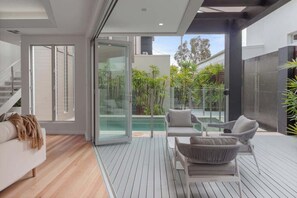 A modern interior with a wooden floor, a sofa, a dining table, and a glass door leading to an outdoor area with plants. - Modern Noosa River Retreat with Heated Pool (Noosa Region)