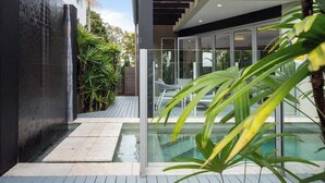 Un área de piscina exterior moderna con cerramiento de vidrio, terraza de madera y abundante vegetación.