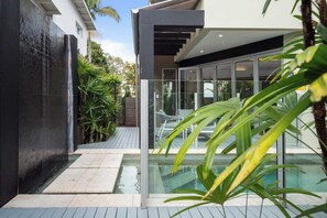 A modern outdoor pool area with a glass enclosure, a wooden deck, and lush greenery. - Modern Noosa River Retreat with Heated Pool (Noosa Region)