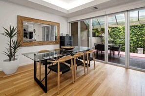 A modern dining area with a glass table, wooden chairs, a large mirror, and a sliding glass door leading to an outdoor patio. - Modern, Airy Duplex Steps to River w/ Heated Pool (Noosa Region)