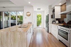A modern kitchen with a central island, stainless steel appliances, and a view to a green outdoor area. - Modern, Airy Duplex Steps to River w/ Heated Pool (Noosa Region)