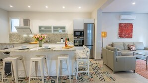 A modern kitchen with a central island, bar stools, a dining area with a sofa, and a patterned rug.