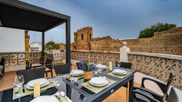 An outdoor dining area with a view of ancient ruins, a grill, and a table set for two.