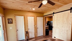 A room with a bed, a ceiling fan, a wooden floor, and a kitchen area visible through an open door. - Brand New! Bright, Airy Cabin Boarding Winslow Hill Elk Viewing Area! (Benezette)