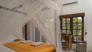 A bedroom with a bed, a table, and a chair, a window with a view of greenery, and a small heater. - Wild Edge Panama - Kumana Safari Lodge (Panama)