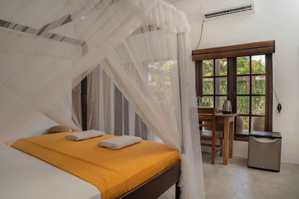 A bedroom with a bed, a table, and a chair, a window with a view of greenery, and a small heater.