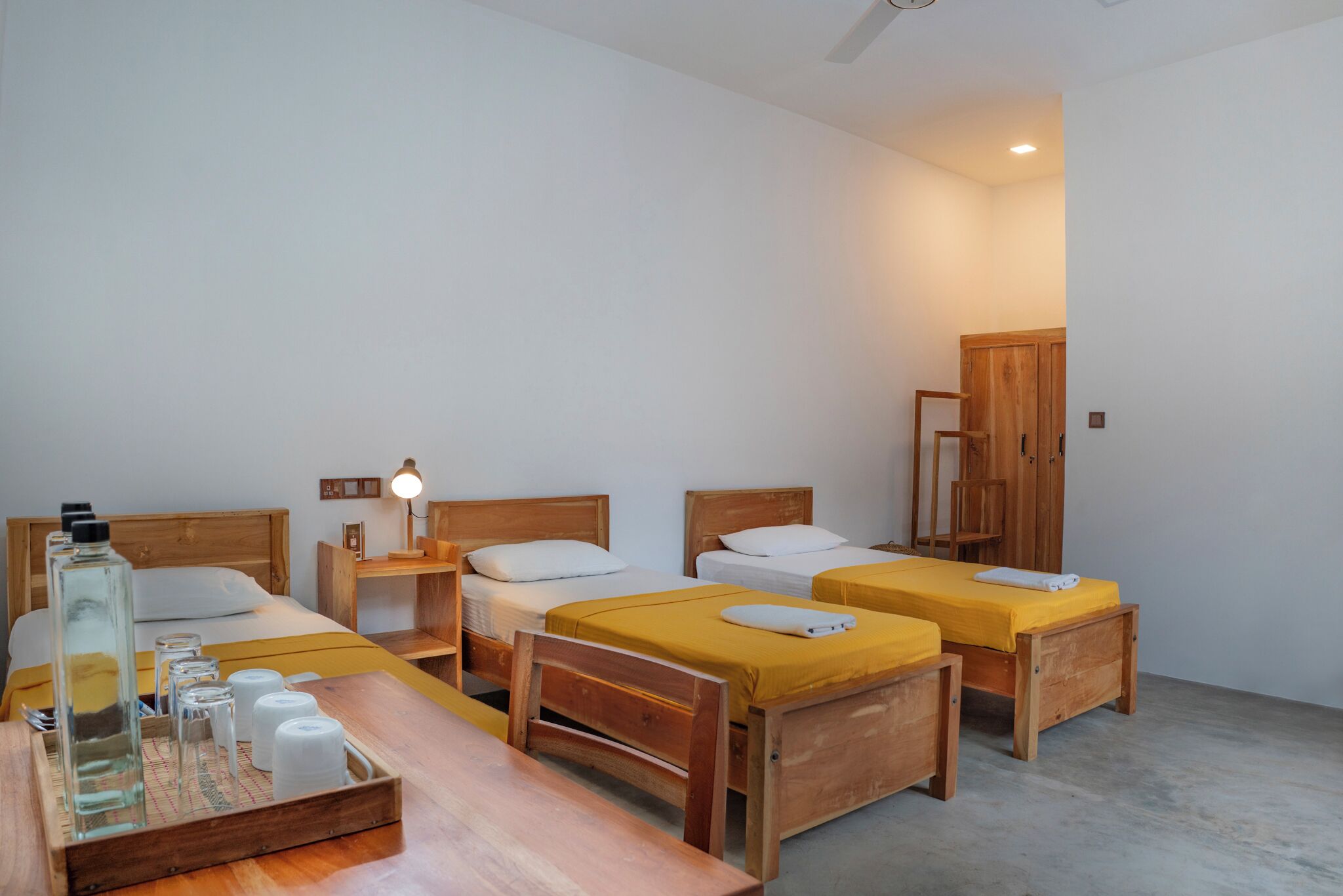 Una habitación con tres camas de madera, un escritorio de madera, un armario de madera y una mesita de noche con una lámpara.