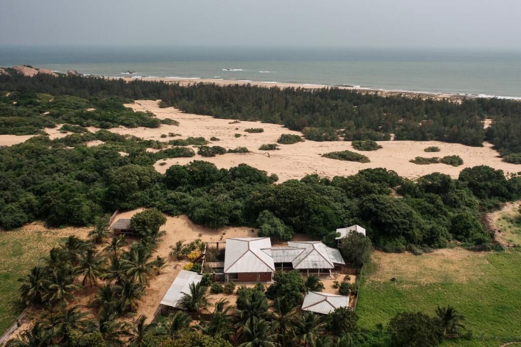 Vista aérea de una propiedad costera con playa, bosque denso y algunos edificios con techos de metal.
