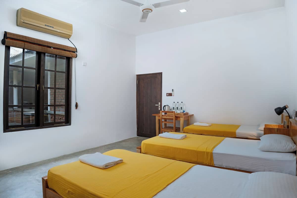 A room with three beds, a desk, a chair, a wardrobe, and a window with a view of a brick building.