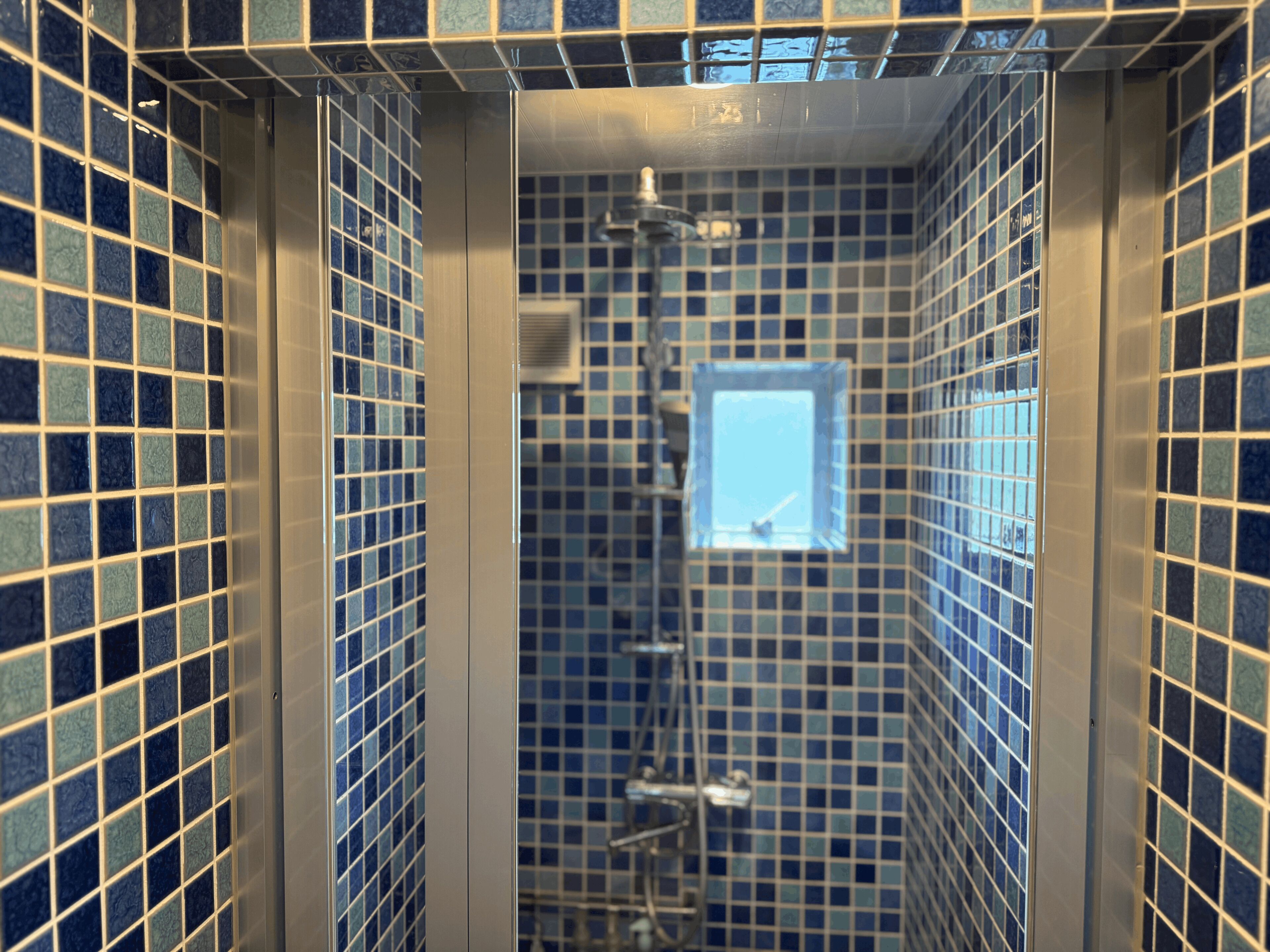 A bathroom with blue mosaic tiles, a shower, and a window.