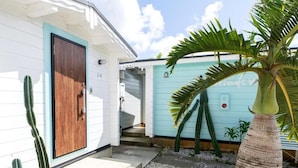 A house with a wooden door, a staircase leading to a covered porch, and a small garden with a palm tree and cacti.